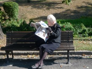 Ältere Frau liest Zeitung auf einer Parkbank.
